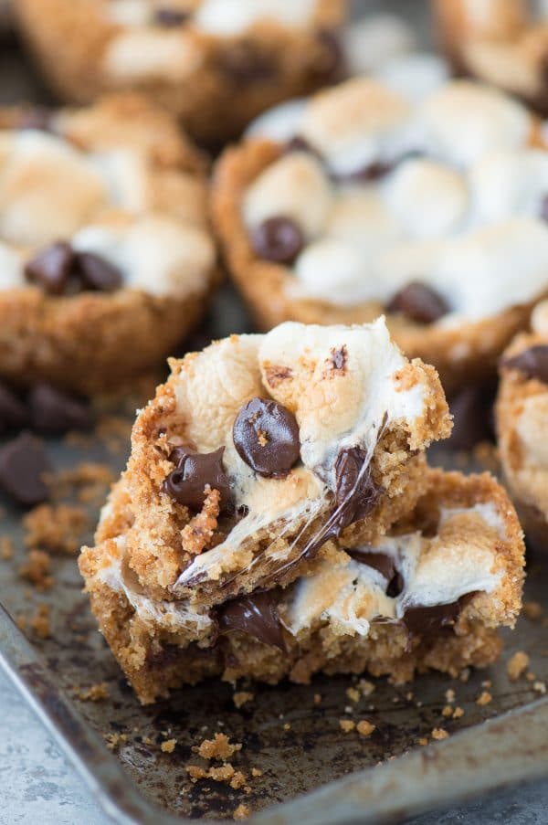 5 ingredient indoor S’mores Cups! Make these in a regular muffin pan. Like a cross between s'mores cookies and s'mores cupcakes! Two half eaten delicious indoor S’mores Cups surrounded by other homemade smores cups on a baking dish.