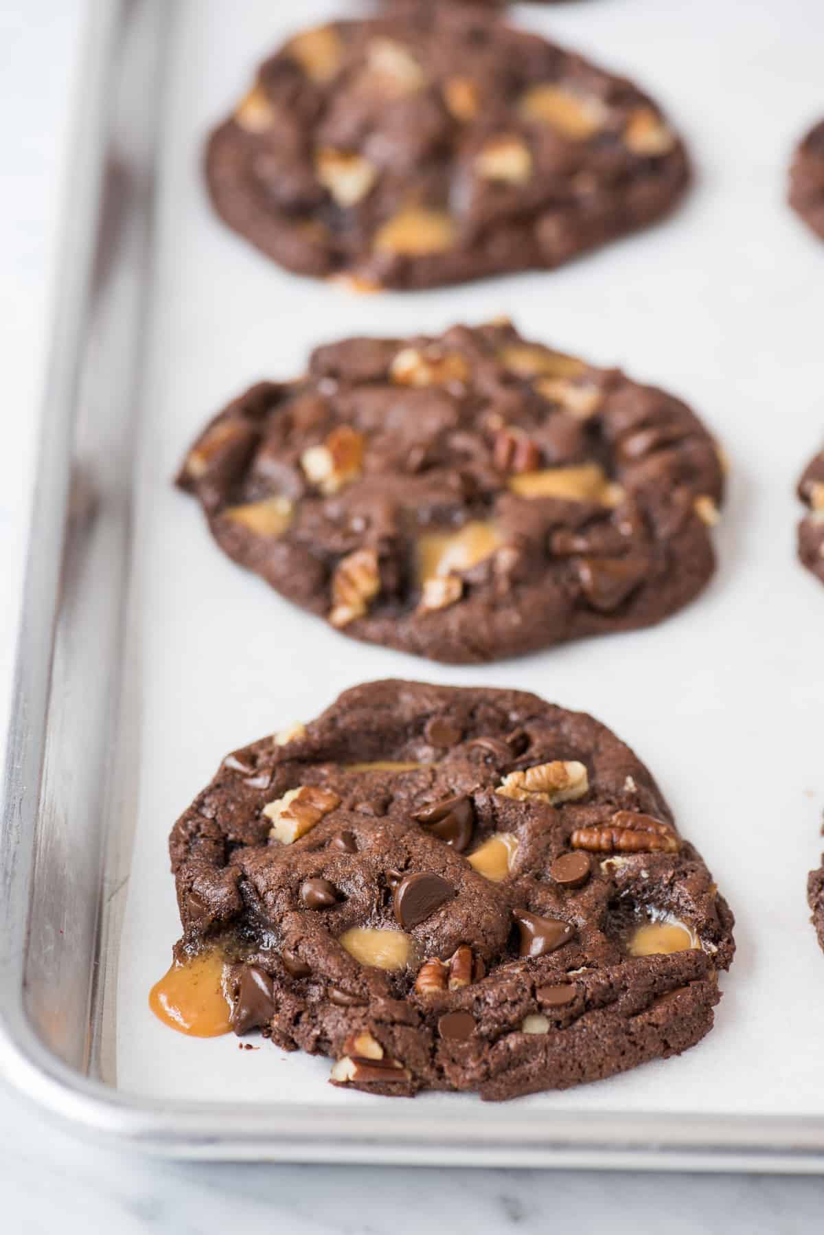 chocolate turtle cookies with caramel, pecans and chocolate chips on white parchment paper on metal baking sheet