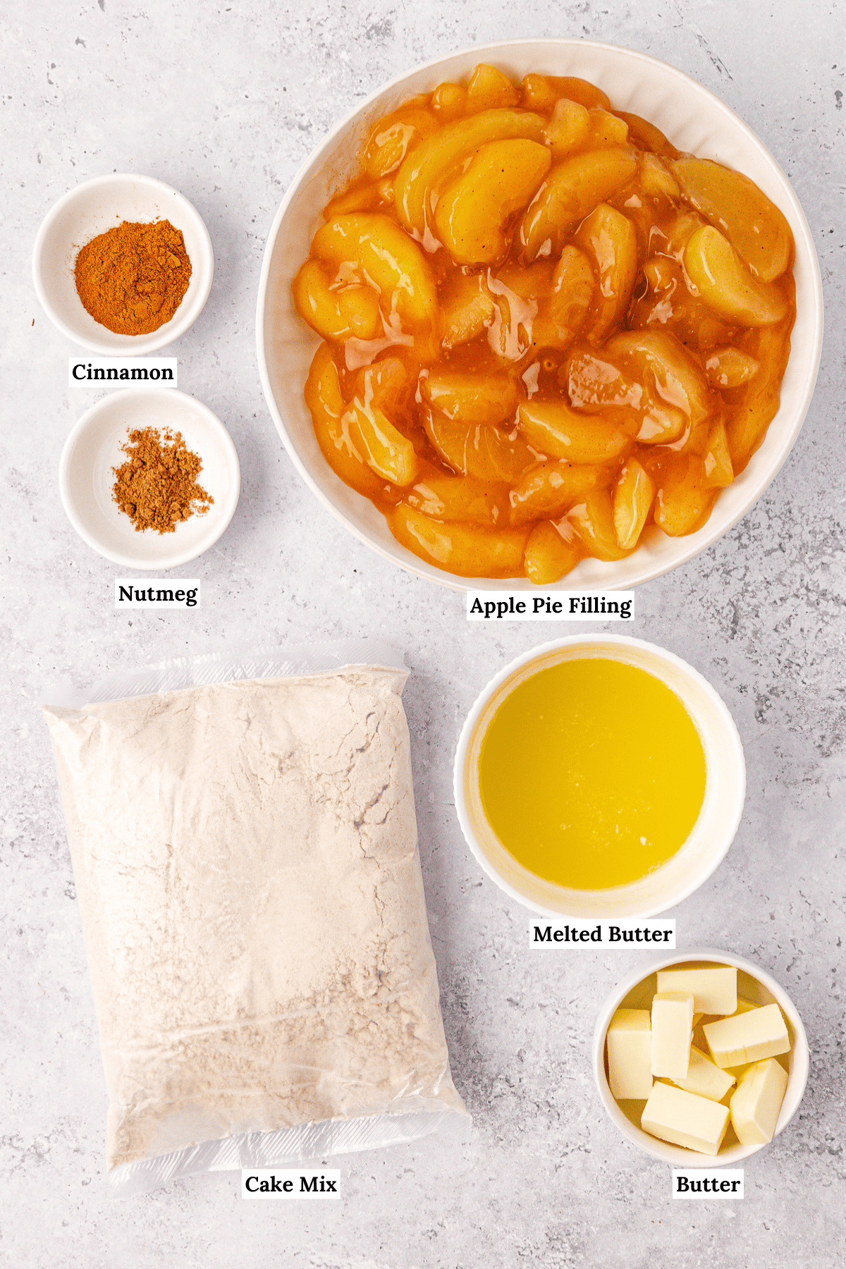 Top view of ingredients for an apple dump cake: a bowl of apple pie filling, small bowls of cinnamon and nutmeg, melted butter in a bowl, a bag of cake mix, and a bowl of sliced butter, all labeled on a light surface.