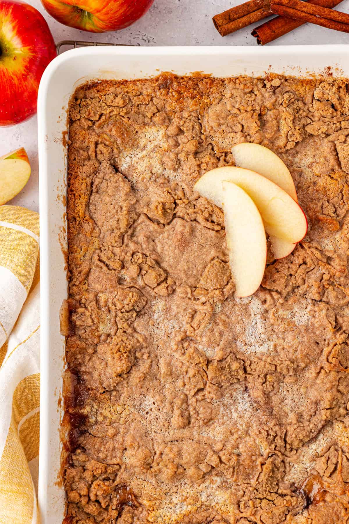 A baking dish filled with a delightful apple crisp topped with three apple slices, resembling an apple dump cake. The dessert boasts a crumbly, golden-brown texture. Surrounding the dish are whole apples, cinnamon sticks, and a yellow-striped towel.