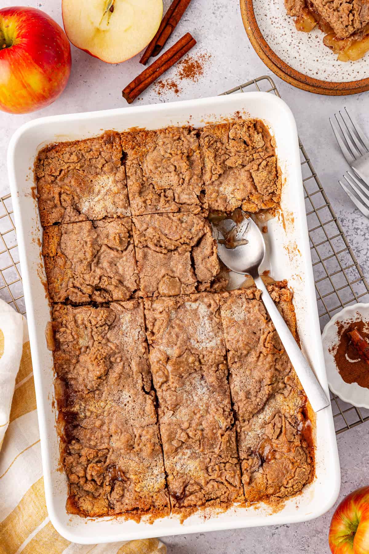 A top view of a baked apple crisp, reminiscent of an apple dump cake, in a rectangular white dish, partially served with a metal spoon. Fresh apples, cinnamon sticks, a plate with a portion of crisp, and a small bowl of cinnamon lie around on the light surface.