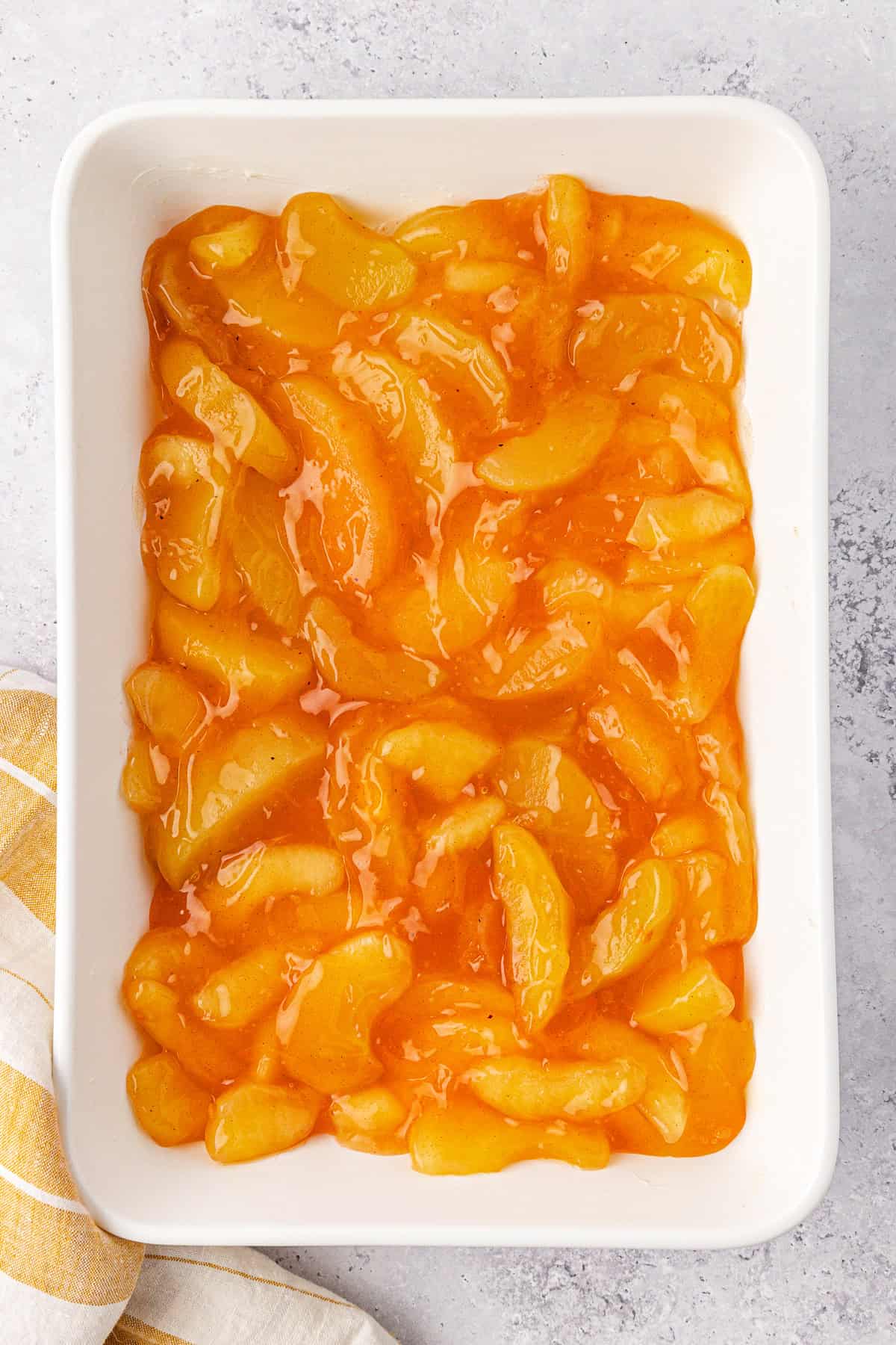 A white baking dish filled with glossy, golden-brown apple pie filling. A folded striped yellow and white cloth napkin is partially visible on the bottom left corner, resting against a light gray surface background.