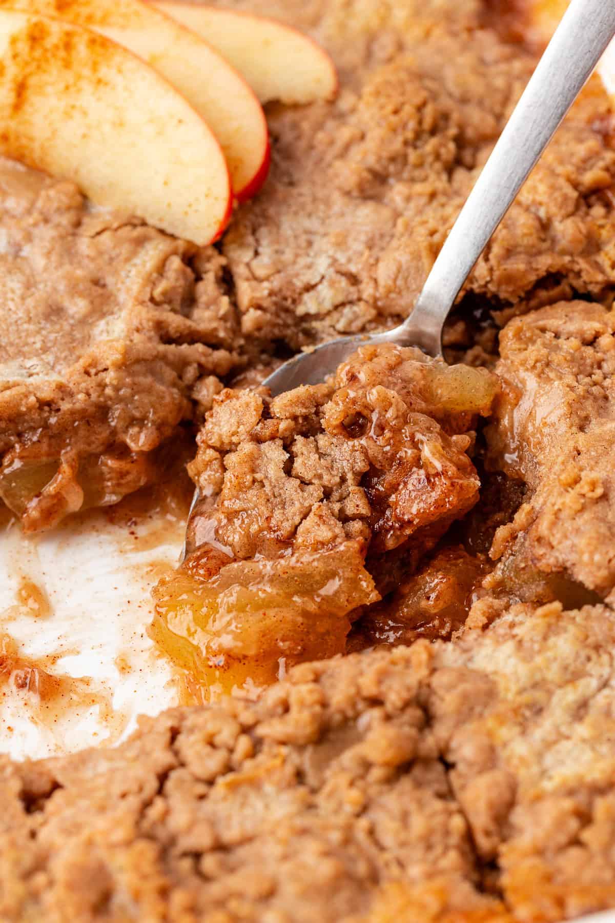 Close-up of a apple pie dump cake with a flaky, golden-brown crust, reminiscent of an apple dump cake. A spoon scoops a portion revealing the warm, gooey apple filling. Slices of fresh red apples garnish the pie in the background.