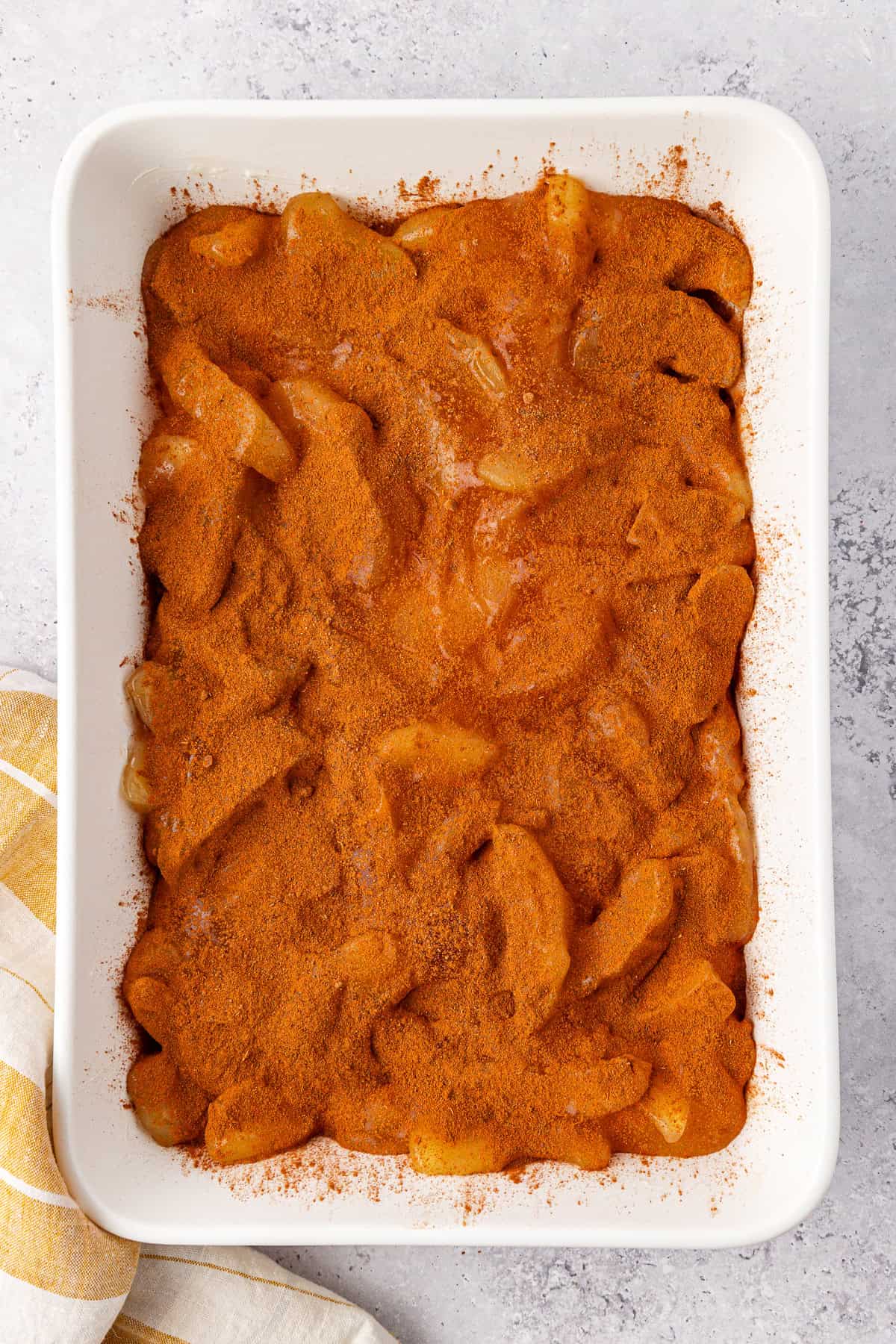 A rectangular baking dish brimming with apple dump cake features sliced apples topped abundantly with cinnamon and sugar, forming a caramelized crust. The dish rests on a light gray surface beside a white and yellow striped cloth.