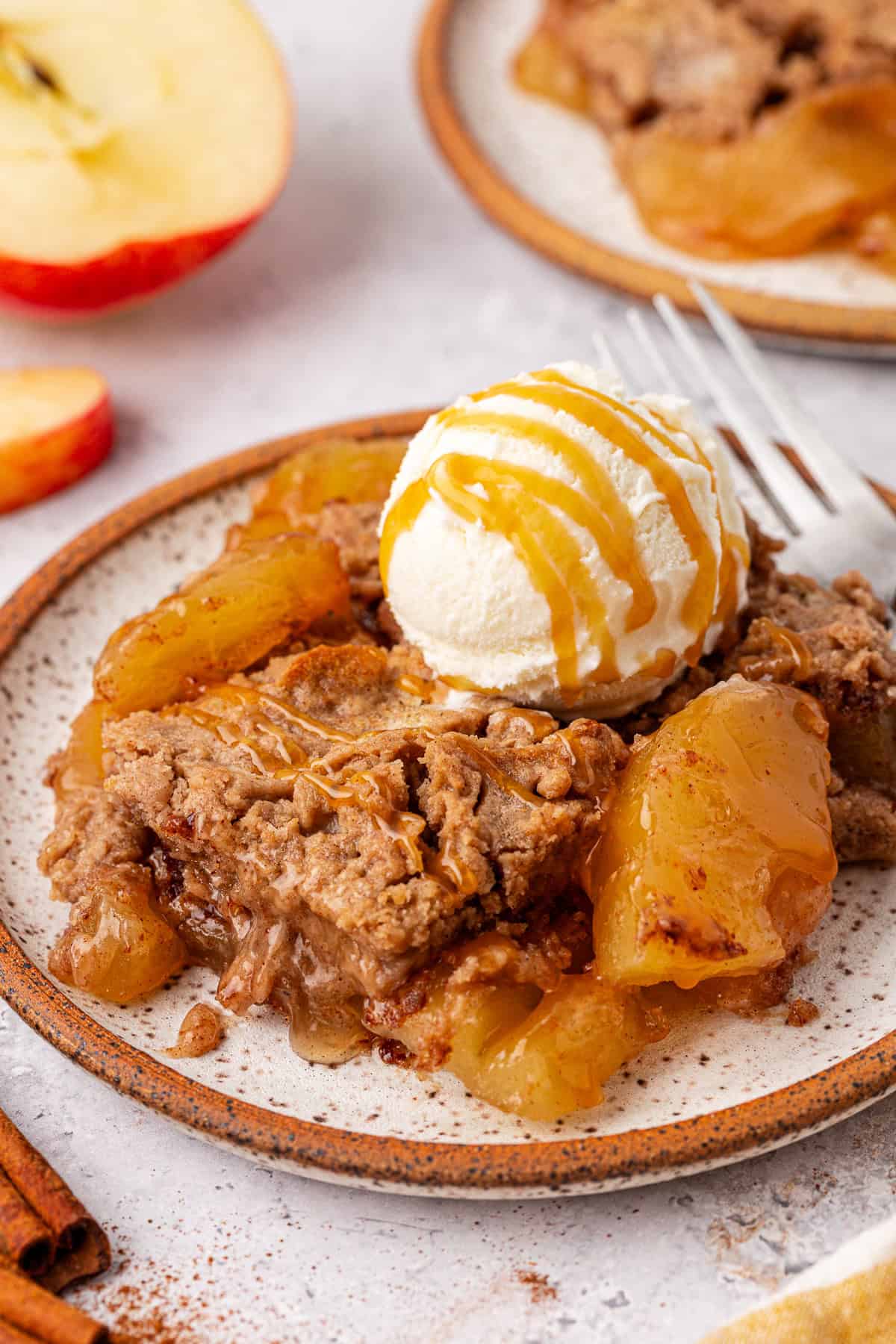 A dessert plate showcasing a warm apple dump cake crowned with a scoop of vanilla ice cream, drizzled in caramel sauce. Slices of apple and a cinnamon stick are artfully arranged in the background on a speckled ceramic plate.