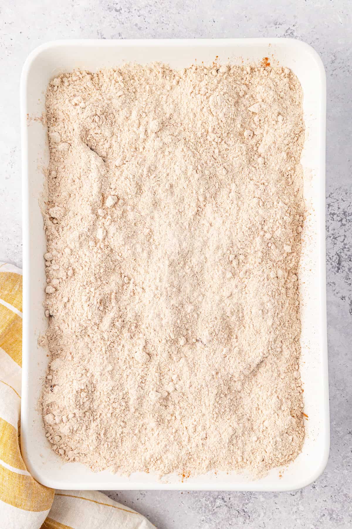 A rectangular white baking dish filled with an even layer of cake mix topping for an apple dump cake, sits on a light gray surface. A yellow and white striped cloth is partially visible beside the dish.