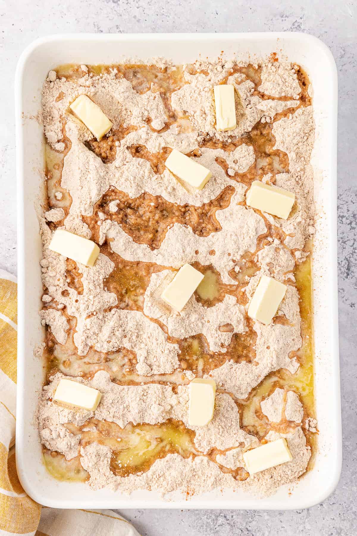 Rectangular baking dish with a apple dump cake before baking. Slices of butter are scattered atop dry cake mix and peaches, reminiscent of an apple dump cake. The dish rests on a textured surface, with a yellow and white cloth draped nearby.