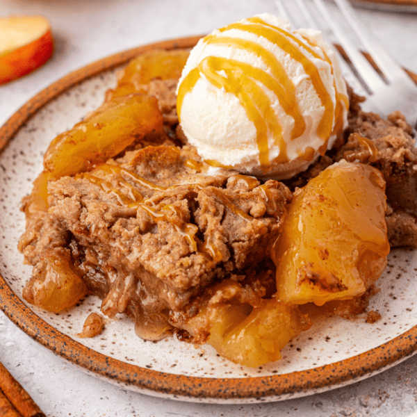 A plate of warm apple crisp, reminiscent of an apple dump cake, is crowned with a scoop of vanilla ice cream drizzled in caramel sauce. The dessert boasts chunks of baked apples beneath a crumbly golden-brown topping. A gleaming fork rests nearby on the ceramic plate.