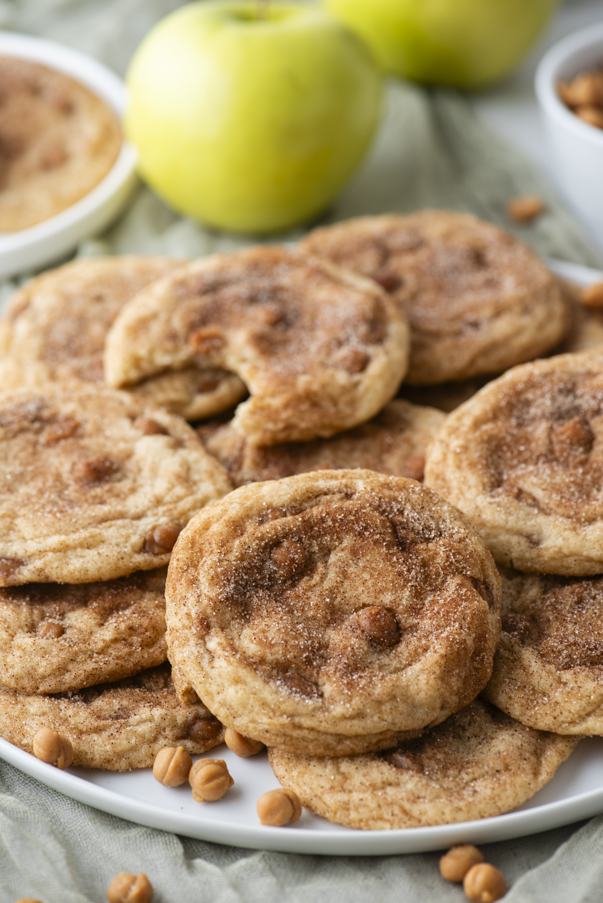 A plate of caramel apple snickerdoodles, packed with cinnamon sugar and caramel chips, some stacked and one with a bite taken out. Green apples and a small bowl of caramel chips are in the background.