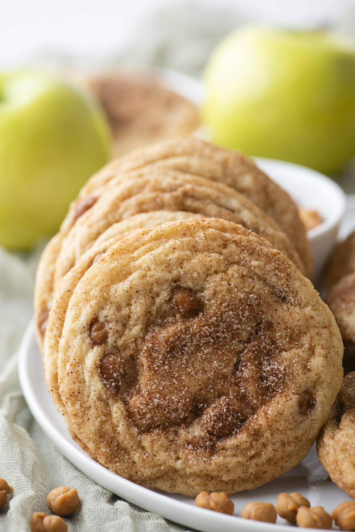 A plate of cinnamon sugar cookies with visible caramel bits sits in front of two green apples on a light cloth background, evoking the cozy flavors of caramel apple snickerdoodles.
