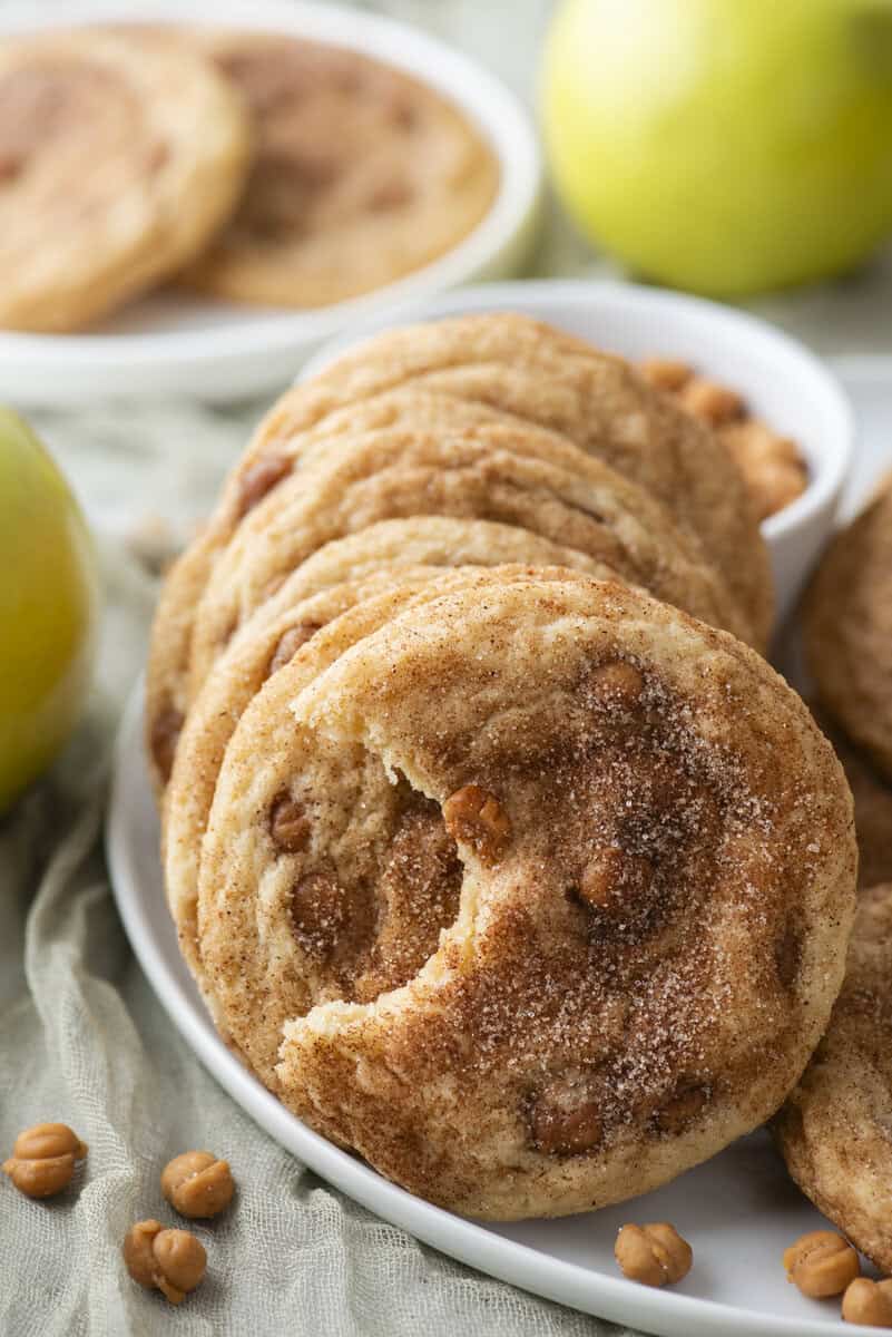 A plate of cinnamon cookies with butterscotch chips, reminiscent of caramel apple snickerdoodles, sits in the foreground—one cookie has a bite taken out. Apples and extra cookies are in the background.