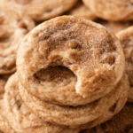 A stack of cinnamon sugar cookies, reminiscent of caramel apple snickerdoodles, with the top cookie showing a bite taken out of it. The cookies have a soft texture and visible sugar and cinnamon sprinkled on top.