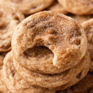 A stack of cinnamon sugar cookies, reminiscent of caramel apple snickerdoodles, with the top cookie showing a bite taken out of it. The cookies have a soft texture and visible sugar and cinnamon sprinkled on top.