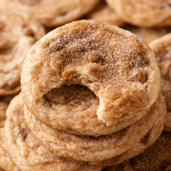 A stack of cinnamon sugar cookies, reminiscent of caramel apple snickerdoodles, with the top cookie showing a bite taken out of it. The cookies have a soft texture and visible sugar and cinnamon sprinkled on top.