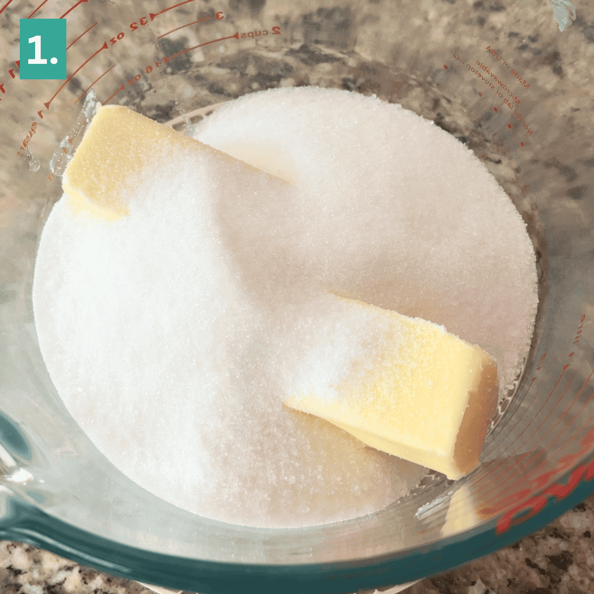 A glass bowl on a counter contains two sticks of butter and a mound of granulated sugar, ready to be mixed for delicious caramel apple snickerdoodles. A green square with the number 1 appears in the top left corner.