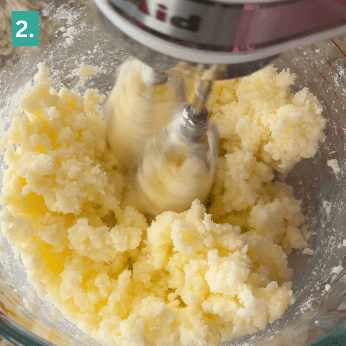 A stand mixer beats butter and sugar in a glass bowl, preparing the base for delicious caramel apple snickerdoodles. The yellow mixture is fluffy, and the mixer blades are in motion. A green square with the number 2 is in the top left corner.