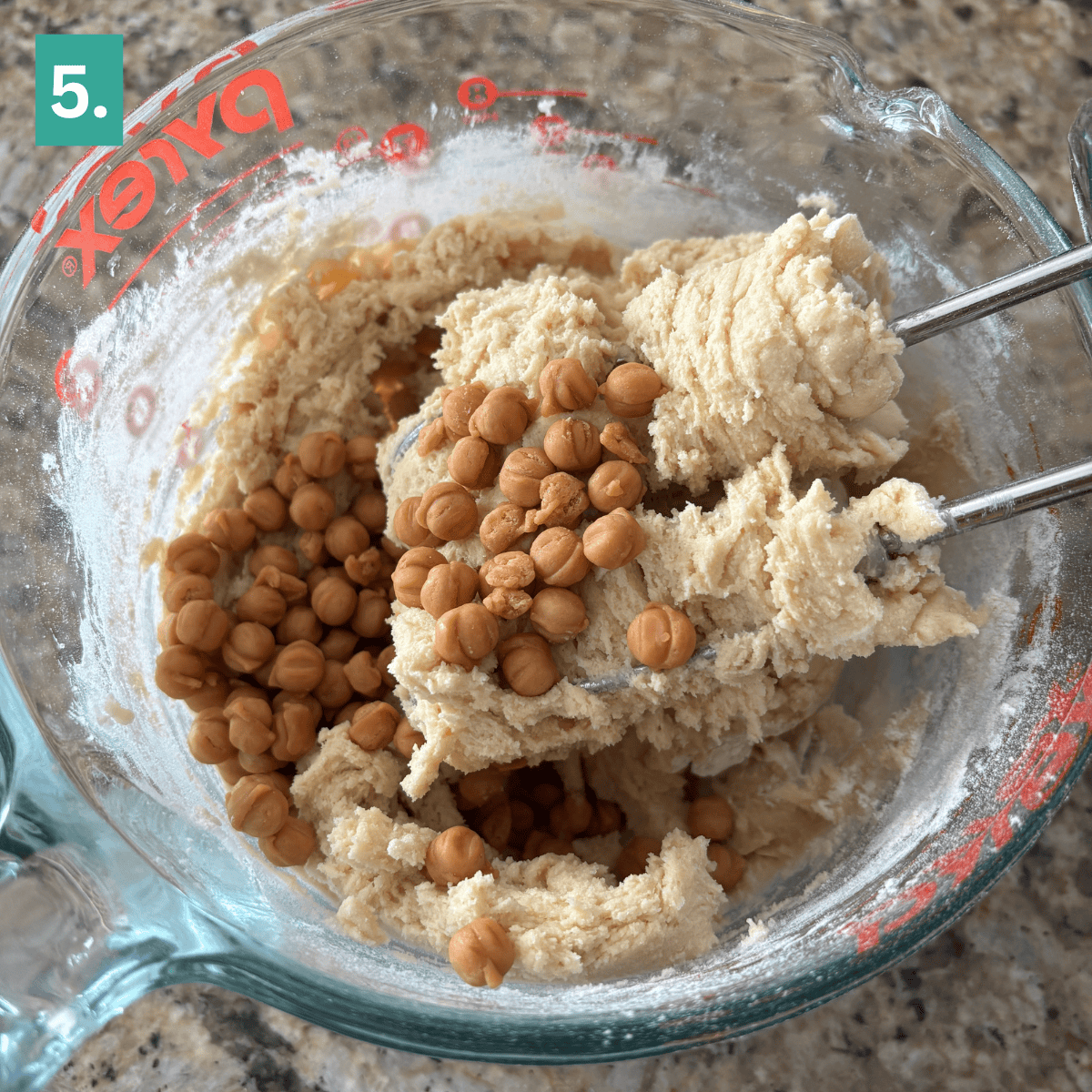 A glass mixing bowl with cookie dough and butterscotch chips, perfect for making Caramel Apple Snickerdoodles. Two metal beaters are in the dough. The bowl sits on a speckled countertop. A green box with the number 5 is in the corner.