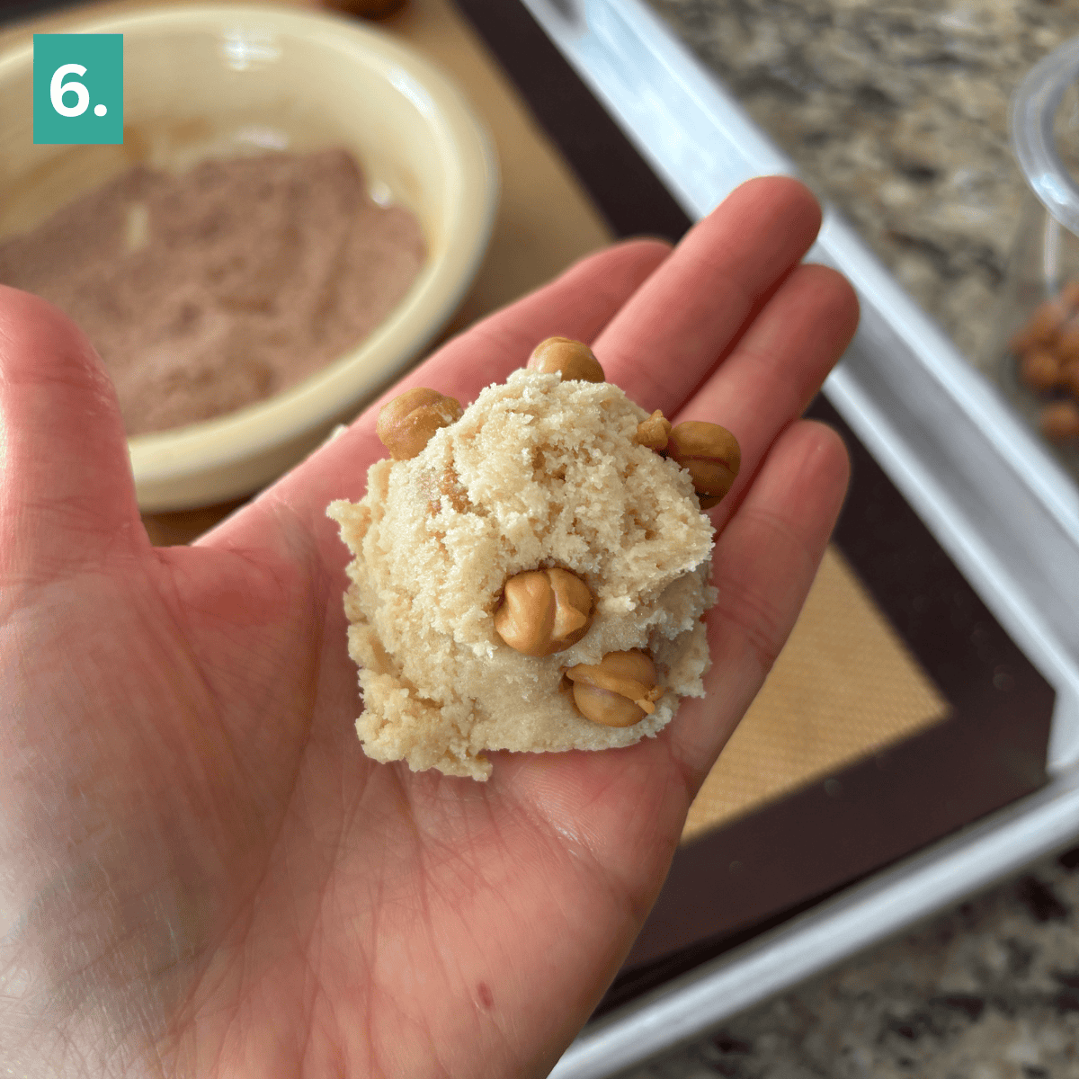 A hand holds a ball of cookie dough with butterscotch chips, ready to be transformed into caramel apple snickerdoodles. In the background are a bowl of cinnamon sugar and a baking tray lined with a silicone mat. A green square with 6. is in the top left corner.