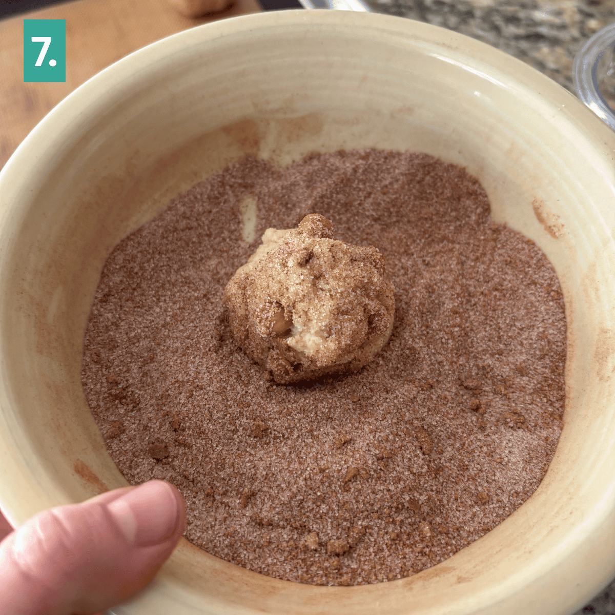 A hand dips a ball of caramel apple snickerdoodle cookie dough into a bowl of cinnamon sugar. The bowl sits on a counter, and a green square with the number 7 is in the top left corner.