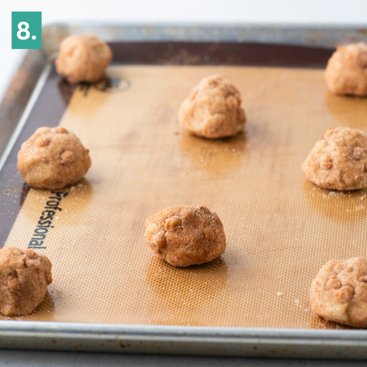 Unbaked caramel apple snickerdoodle cookie dough balls spaced out on a silicone baking mat on a metal baking sheet, ready to go into the oven. A green box with the number 8 is in the top left corner.