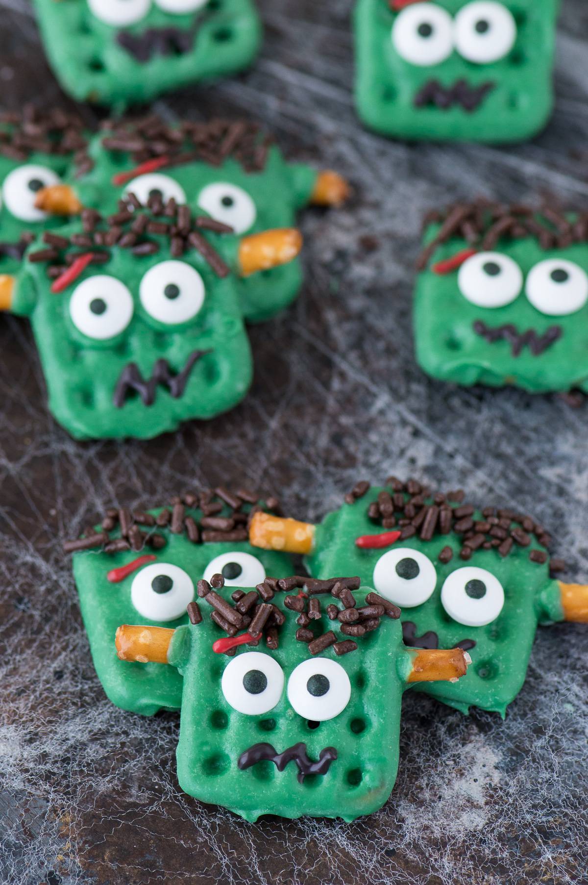 frankenstein treats made out of square waffle pretzels dipped in green chocolate with decorative embellishments on dark background