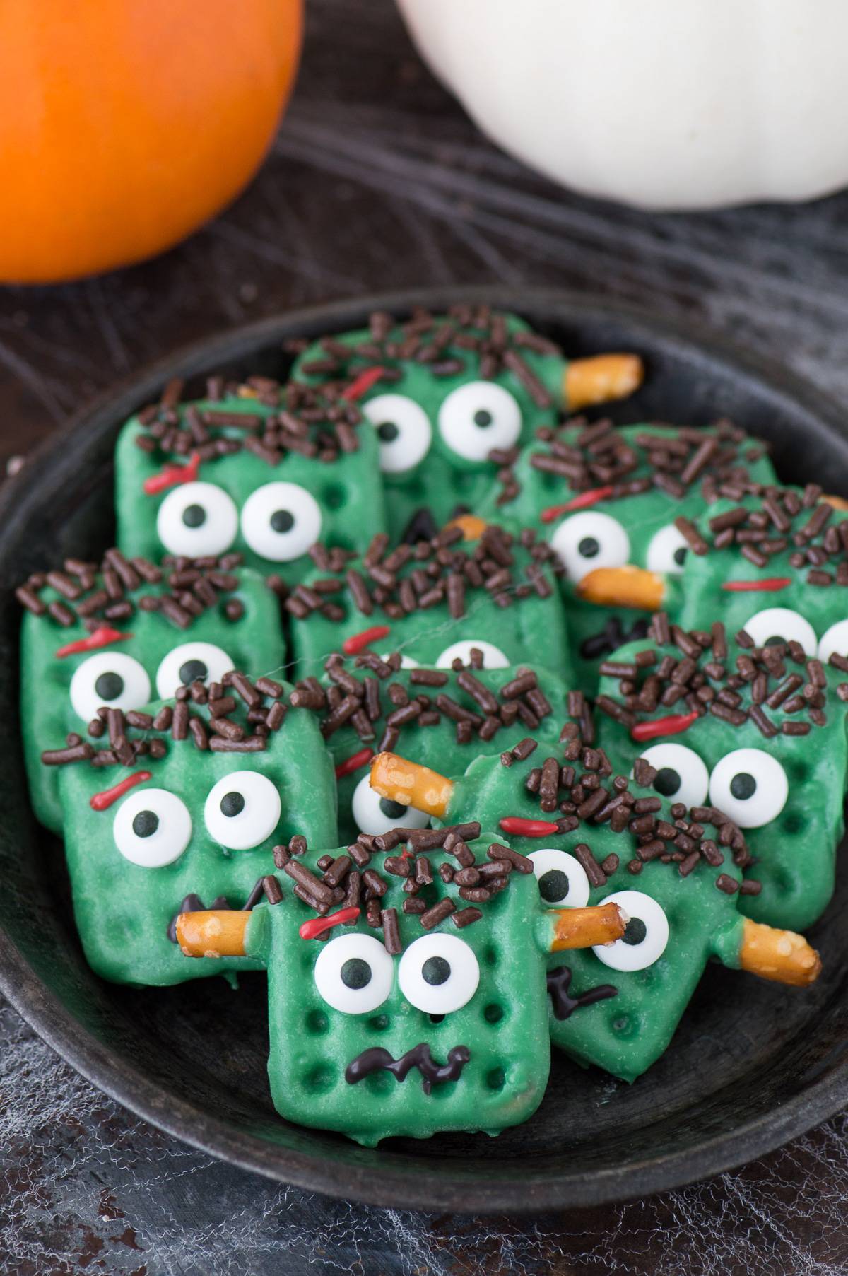 frankenstein treats made out of square waffle pretzels dipped in green chocolate with decorative embellishments on dark plate on dark background