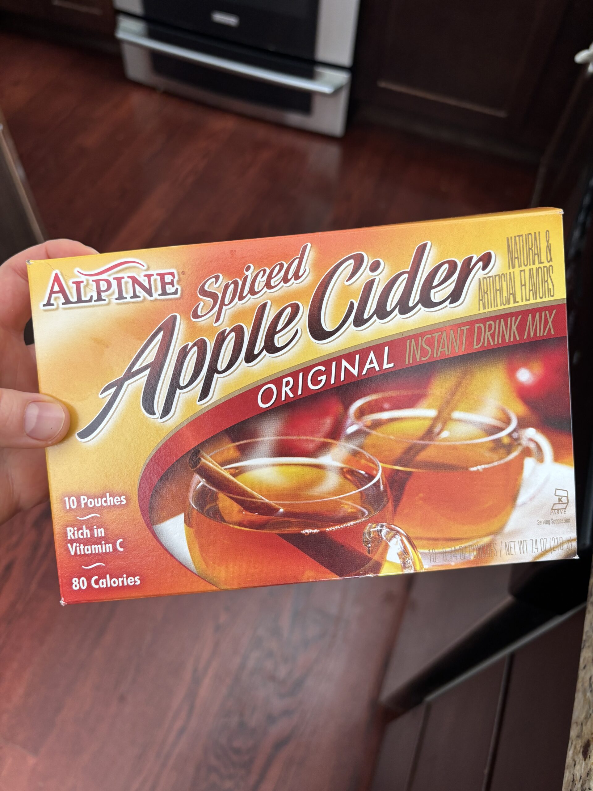 A hand holds a box of Alpine Spiced Apple Cider Original Instant Drink Mix with images of two mugs of cider and cinnamon sticks on the front. The box states it contains 10 pouches, is rich in vitamin C, and has 80 calories—an inviting choice for sipping alongside freshly baked caramel apple snickerdoodles.