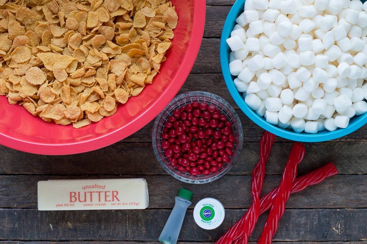 corn flakes in red bowl, marshmallows in blue bowl, red m&ms in clear bowl, stick of butter, green food coloring and twizzler's pull n peels on wood background
