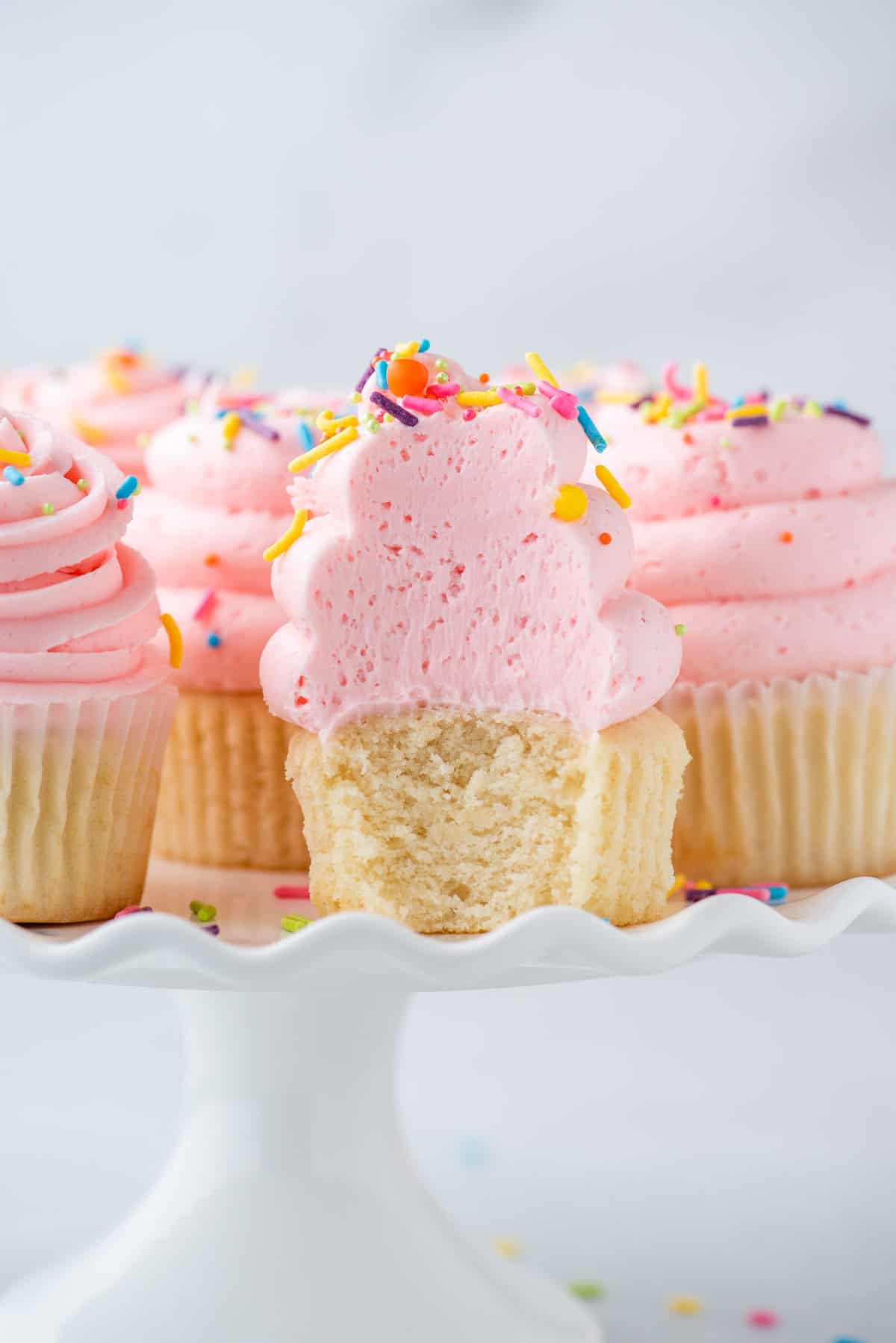vanilla cupcakes with pink frosting and sprinkles with bite removed on white cake stand