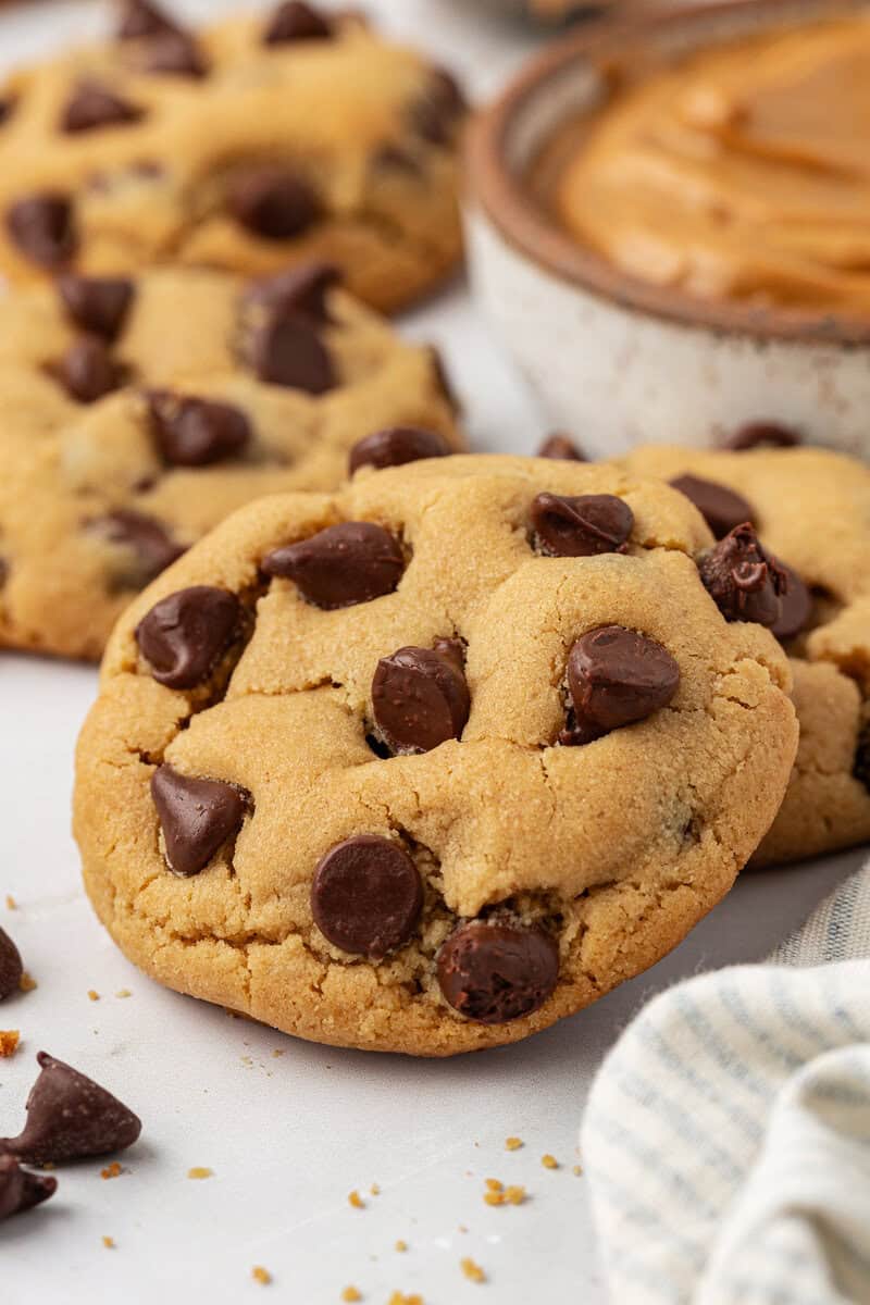Close-up of golden brown peanut butter cookies studded with large chocolate chips on a white surface, with a bowl of creamy peanut butter and a striped cloth in the background.