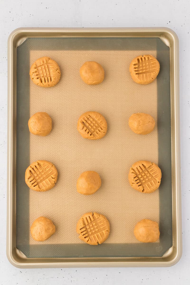 A baking sheet lined with a silicone mat holds ten round balls of peanut butter cookie dough, with four of them pressed with a fork to create the classic crisscross pattern of peanut butter cookies.