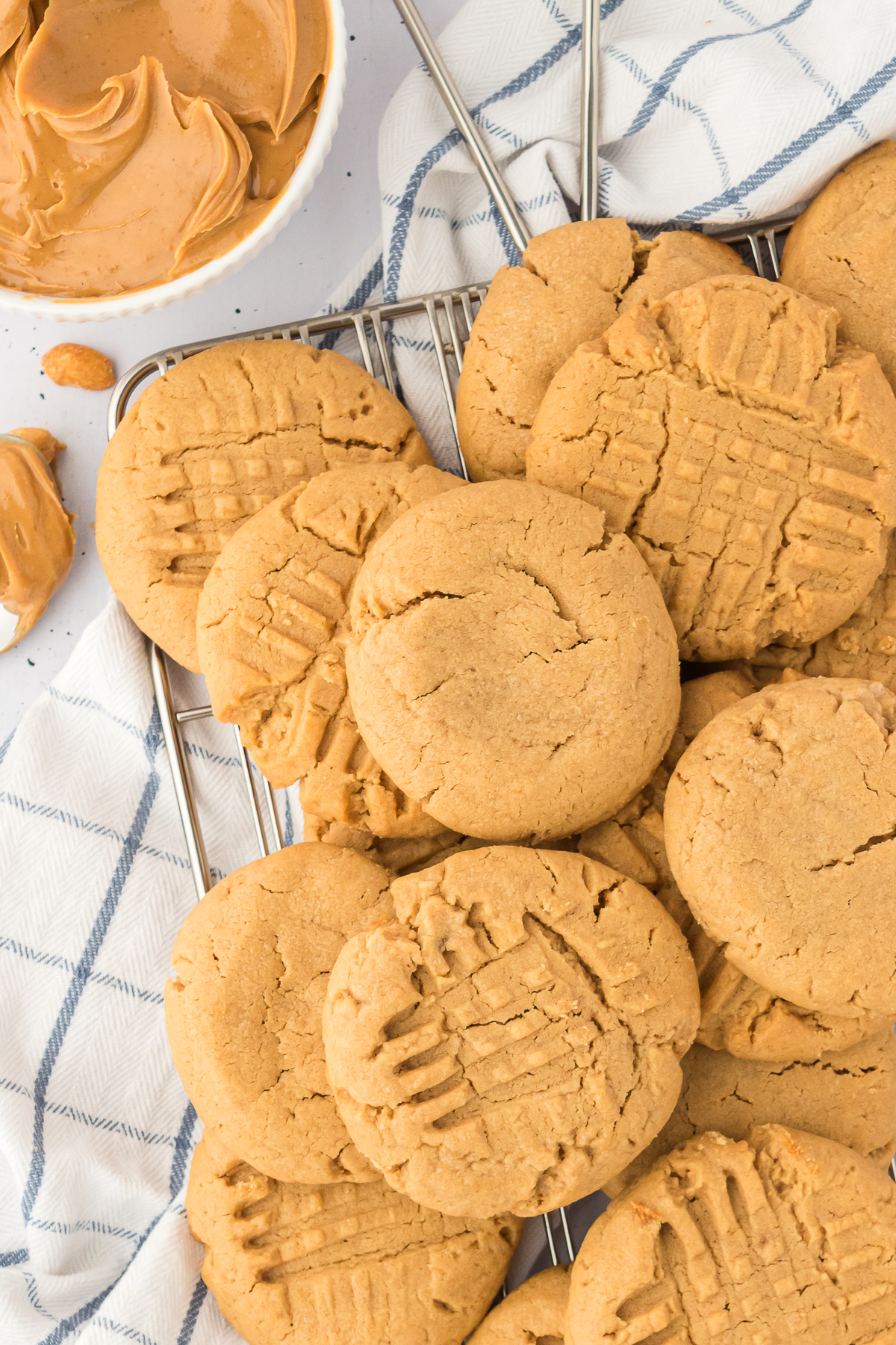 A cooling rack piled with classic peanut butter cookies, each marked with a crisscross fork pattern, sits next to a bowl of creamy peanut butter on a white and blue checkered cloth. Perfect for peanut butter cookie lovers.