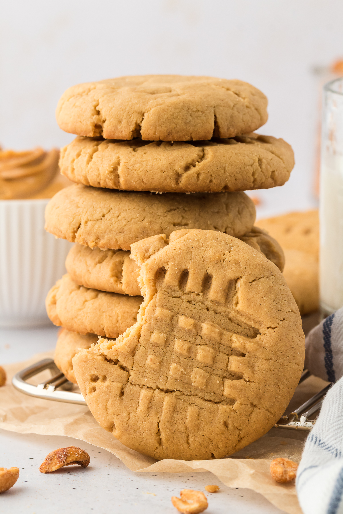 A stack of peanut butter cookies sits on parchment paper, with one in front showing a large bite and the classic crisscross fork pattern. Scattered peanuts and a glass of milk complete this peanut butter cookie scene.