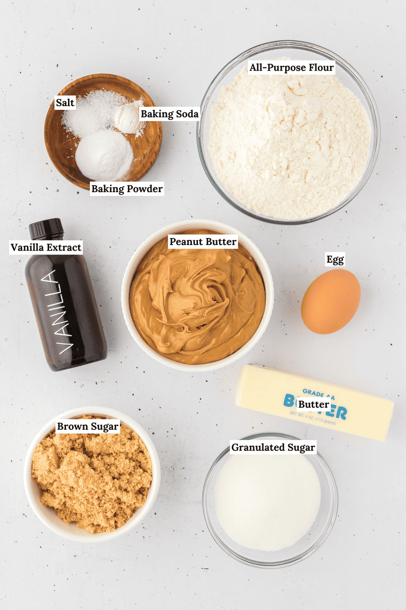 Top-down view of labeled baking ingredients for peanut butter cookies on a white surface: all-purpose flour, salt, baking soda, baking powder, vanilla extract, peanut butter, egg, brown sugar, granulated sugar, and a stick of butter.