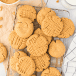 A cooling rack holds several golden-brown peanut butter cookies, some with classic crisscross fork marks. Nearby are a glass of milk, a striped cloth, and part of a bowl brimming with peanut butter cookie dough.