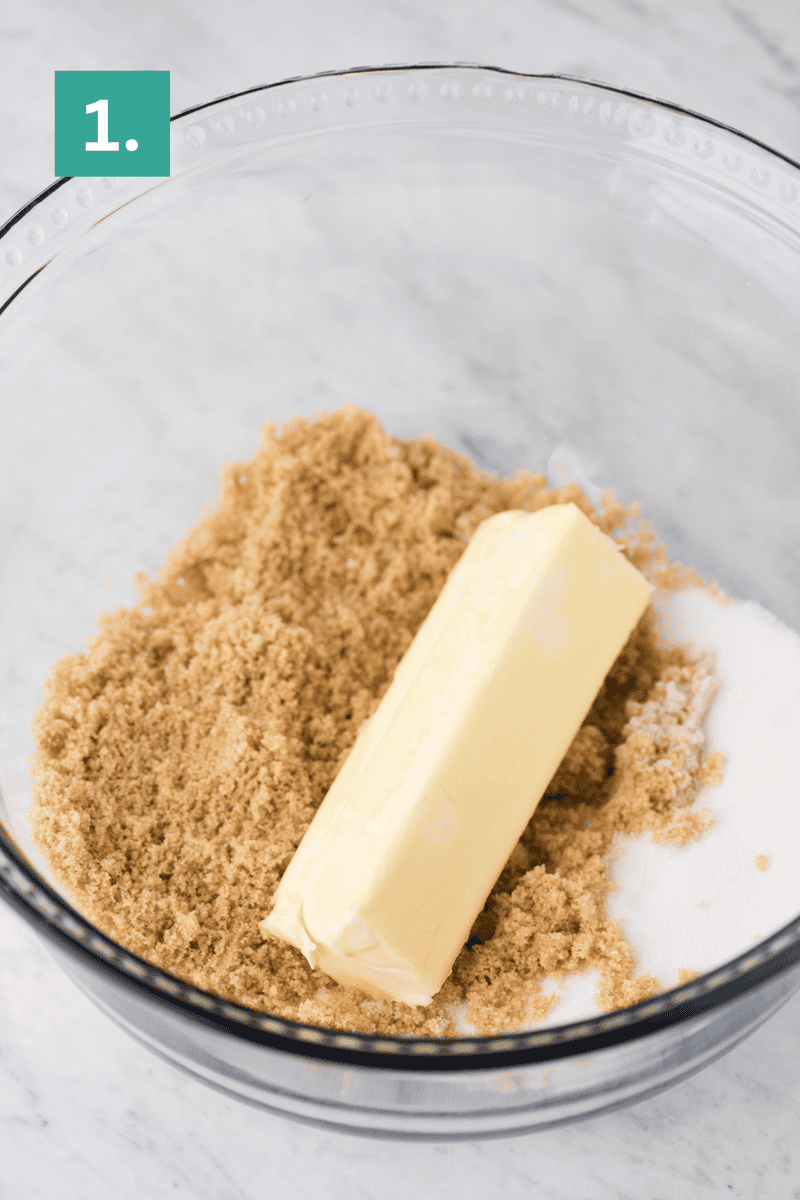 A glass bowl containing a stick of butter, brown sugar, and white sugar sits on a marble surface. A green box with the number 1 is in the top left corner, marking step one of delicious peanut butter cookies preparation.
