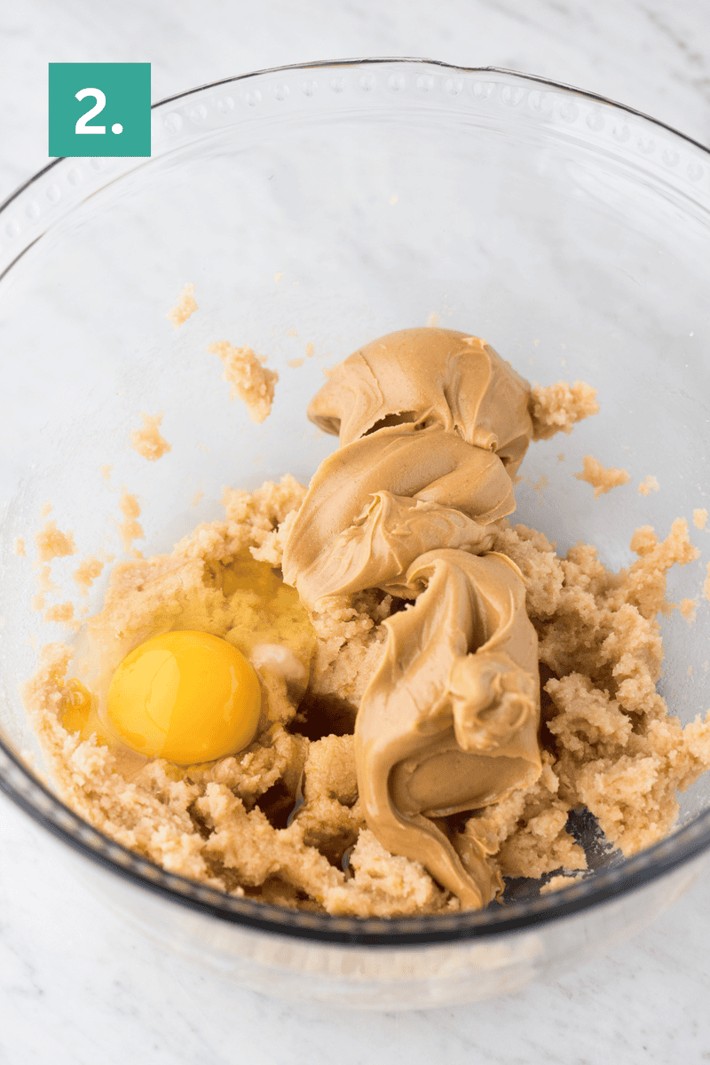 A glass bowl containing brown sugar, an egg, vanilla extract, and dollops of peanut butter for peanut butter cookies sits on a marble countertop. A green label with the number 2 is in the top left corner.
