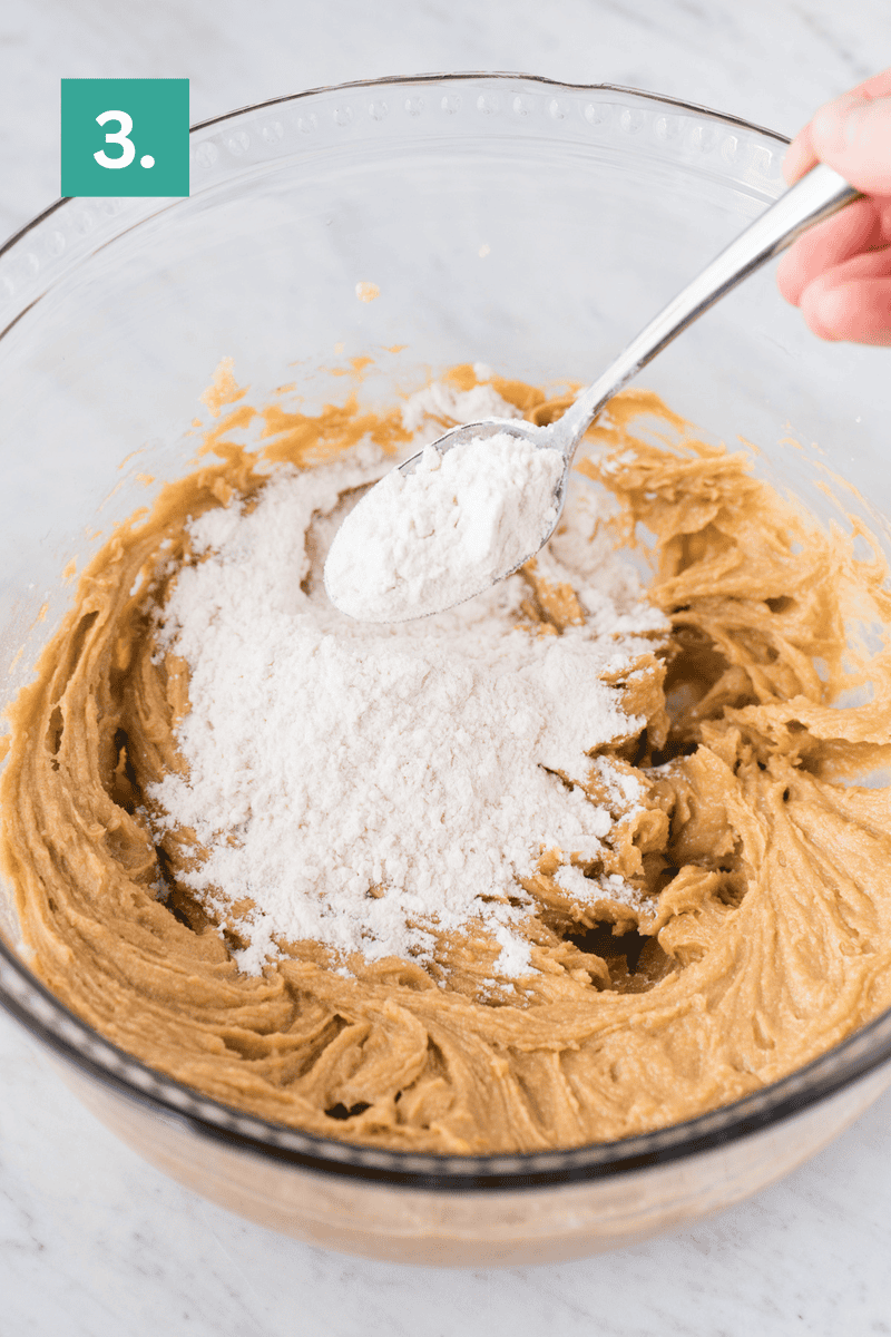 A spoon adds flour to a bowl of light brown peanut butter cookie dough. Step 3 is shown in a green box in the top left corner. The bowl sits on a white marble surface.