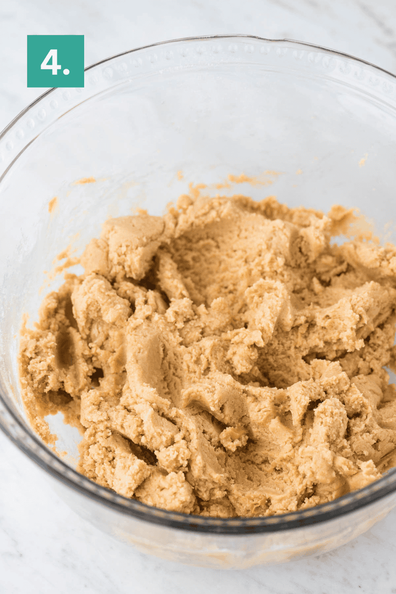 A clear glass bowl filled with creamy, mixed peanut butter cookie dough sits on a marble surface. In the top left corner, a green square with the number 4. indicates a step in a recipe.