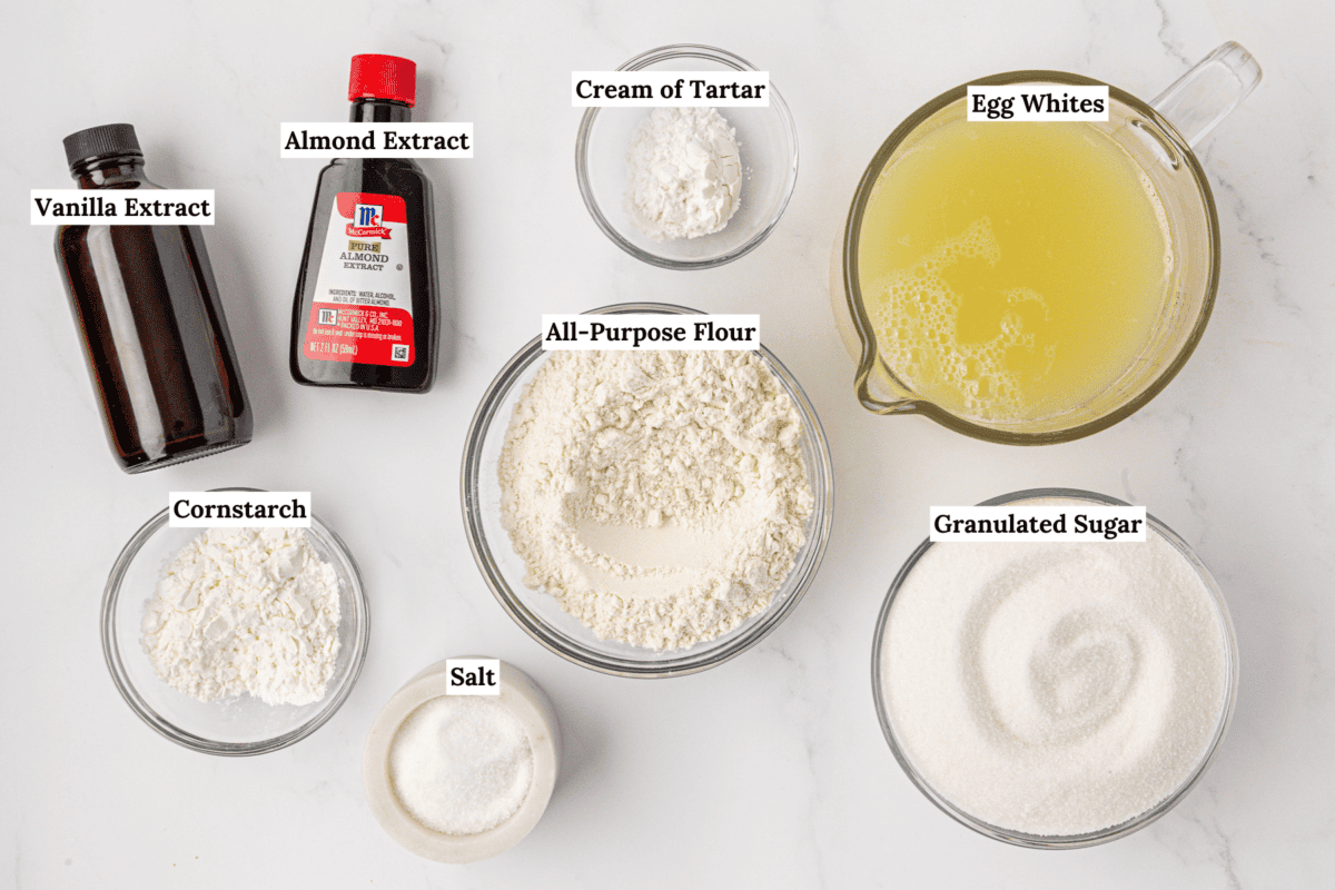 Labeled ingredients for baking angel food cake on a white surface: vanilla extract, almond extract, cream of tartar, egg whites, all-purpose flour, cornstarch, salt, and granulated sugar in bowls and containers.