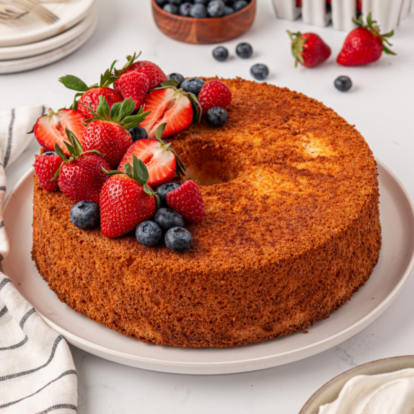 A golden-brown angel food cake topped with fresh strawberries and blueberries sits on a white plate, surrounded by more berries and a striped cloth napkin on a white table.