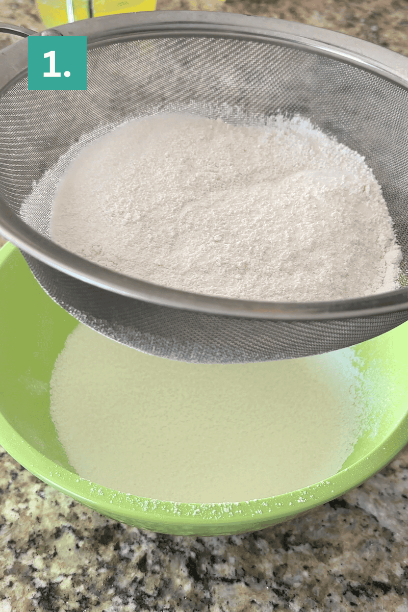 A metal sieve sifting white flour into a green bowl on a granite countertop&mdash;the first step in making angel food cake. The number 1 in a green box appears in the image corner, marking the beginning of the process.