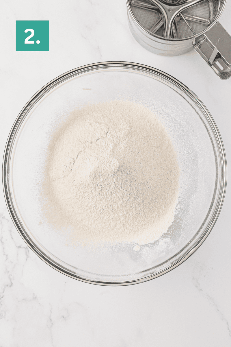 A glass bowl filled with sifted flour, perfect for making angel food cake, sits on a white marble surface. A flour sifter is partially visible in the top right corner, and a green box with the number 2 is in the top left.