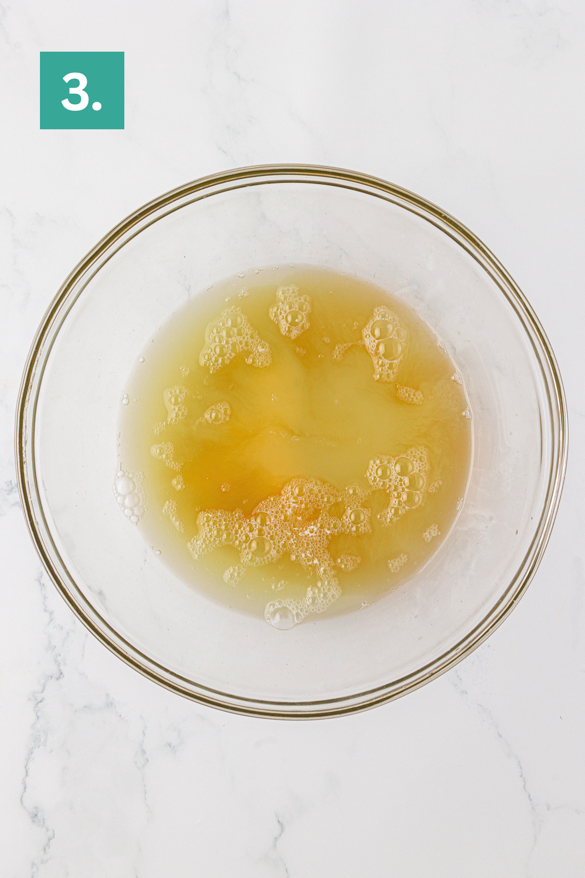 A glass bowl with lightly beaten eggs and bubbles on the surface, ready for angel food cake, sits on a white marble countertop. A green box with the number 3 appears in the upper left corner.