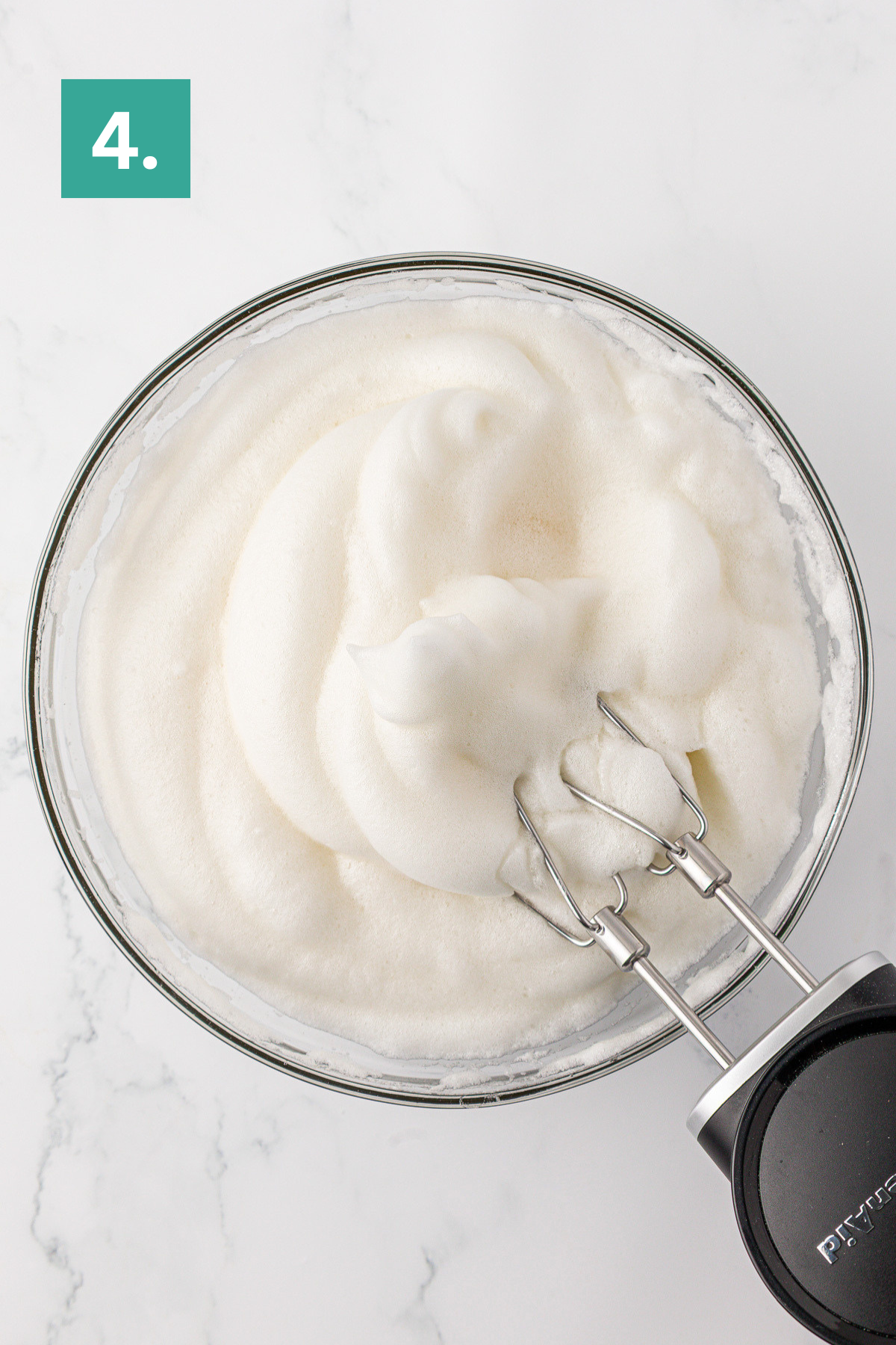 A glass bowl filled with stiffly beaten egg whites for an angel food cake is shown from above, with a hand mixer resting inside. A green box labeled 4. appears in the top left corner.