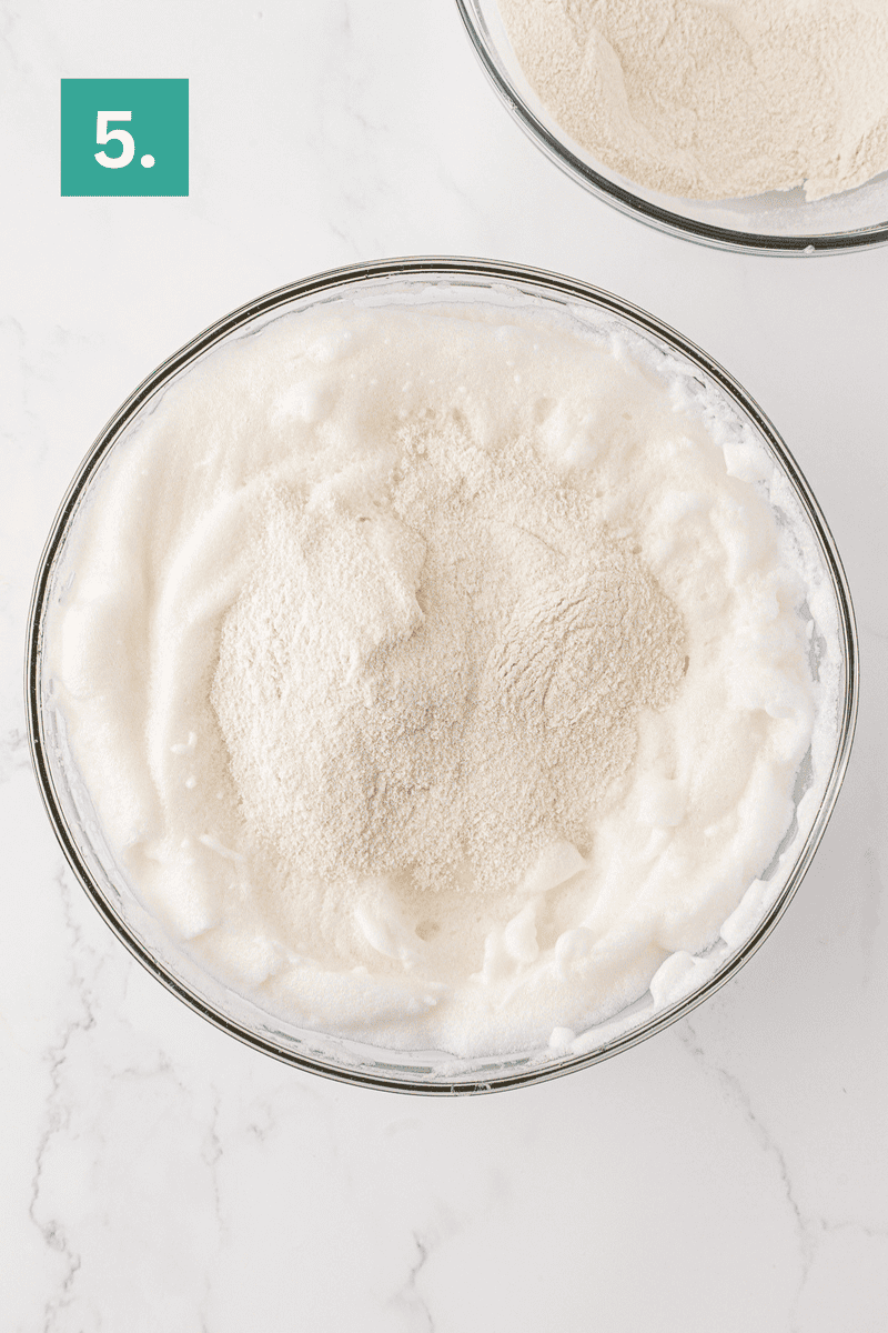 A glass bowl filled with beaten egg whites and a mound of sifted dry ingredients&mdash;perfect for making angel food cake&mdash;sits on a white marble surface. A green box with the number 5 appears in the top left corner.