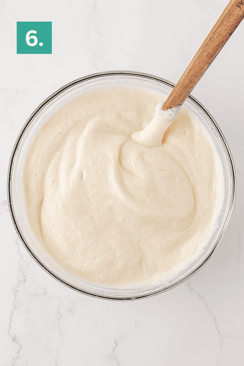 A glass bowl filled with fluffy, whipped cream mixture for angel food cake sits with a wooden spoon on a white marble surface. A green square with the number 6. appears in the top left corner.
