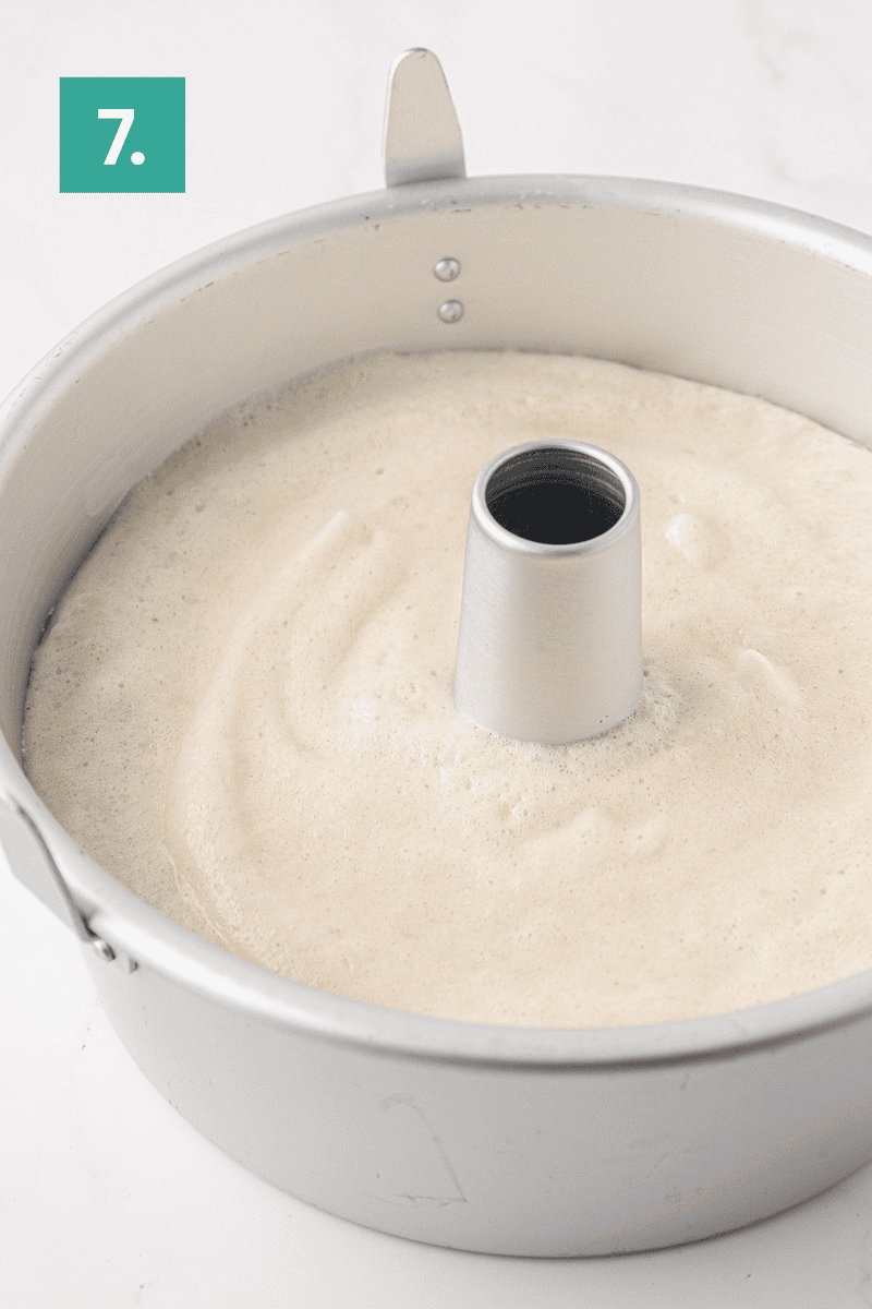 A metal tube pan filled with light, airy angel food cake batter sits on a white surface. A green box with the number 7 appears in the top left corner of the image.
