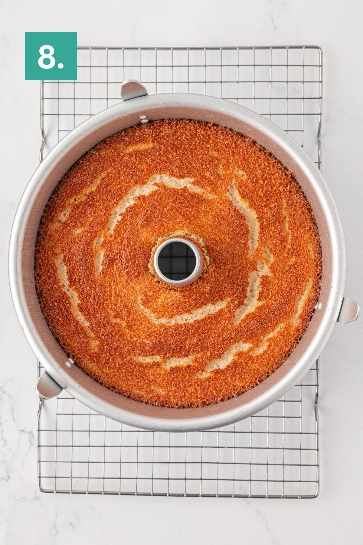 A golden-brown angel food cake in a round tube pan cools on a wire rack; a green label with the number 8 is in the top left corner.