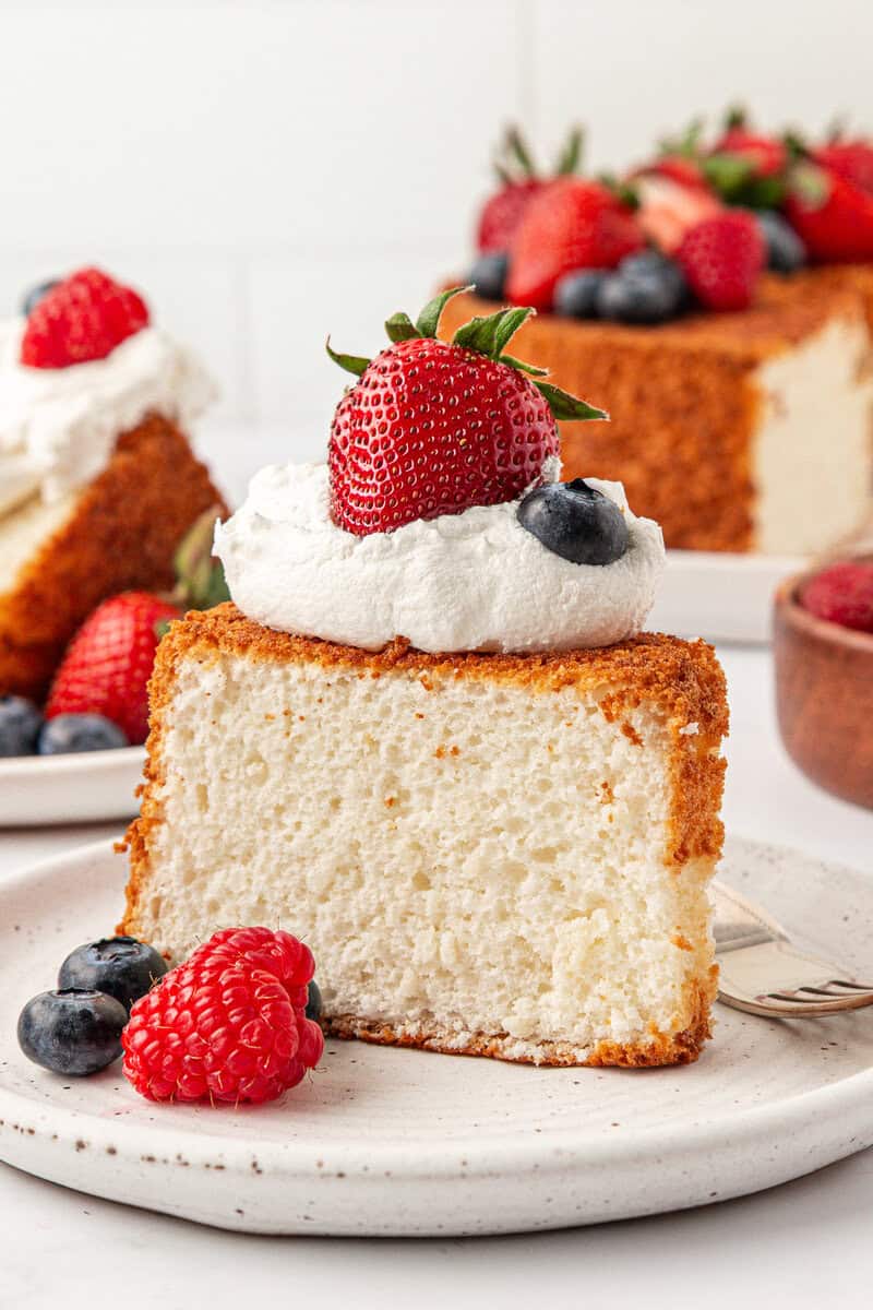 A slice of fluffy angel food cake topped with whipped cream, a strawberry, and a blueberry is served on a white plate with extra raspberries and blueberries. The rest of the angel food cake appears temptingly in the background.